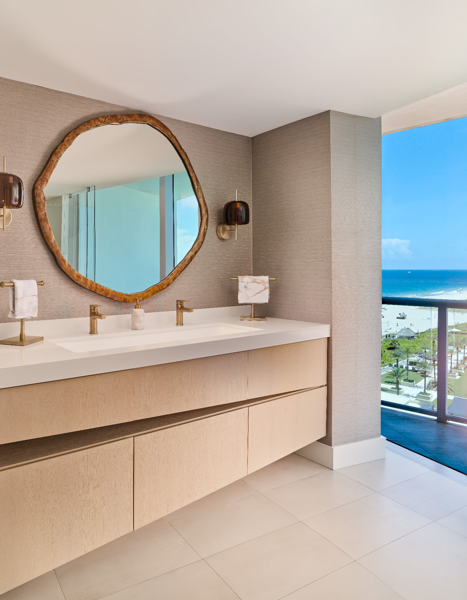 Primary bath with grasscloth walls, floating light-oak vanity, organic-edge mirror and aged brass fittings overlooking the ocean
